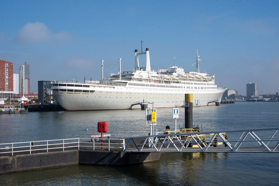 A modern cruise ship docked at the Rotterdam Cruise Terminal with the De Rotterdam towers behind