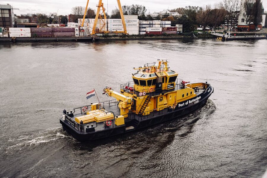 A yellow tugboat working in the Port of Rotterdam with shipping containers stacked behind